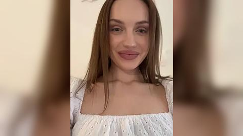 Media: Video of a young woman with fair skin and long brown hair, wearing a white off-shoulder blouse with subtle polka dot detailing, smiling softly. Background is blurred, indoor setting.