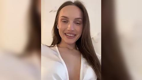 Media: Video of a smiling, light-skinned woman with long brown hair wearing a white, low-cut V-neck blouse. Background is blurred, suggesting an indoor setting with neutral-colored walls.