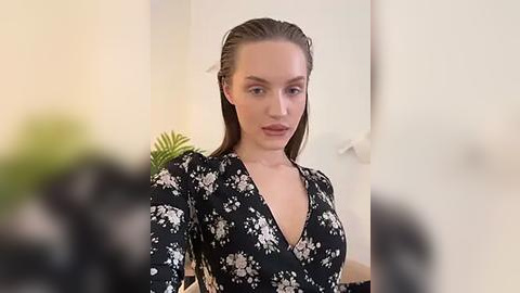 Media: A video of a fair-skinned woman with wet, slicked-back hair, wearing a black floral dress, standing indoors with blurred greenery in the background.