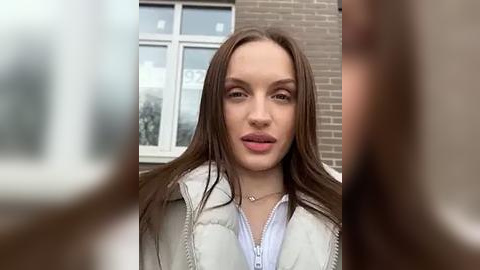 Media: Video of a young Caucasian woman with long brown hair, fair skin, and a light jacket, standing in front of a brick wall with a window. Her expression is neutral.