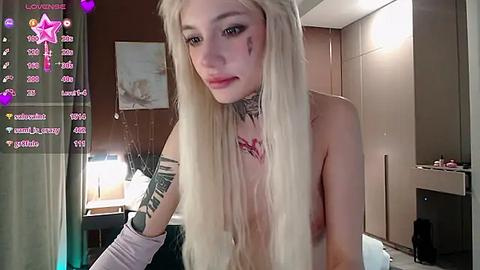 Media: Video of a topless, pale-skinned, blonde woman with a tattooed arm, blood on her face, in a dimly lit bedroom with a TV in the background.