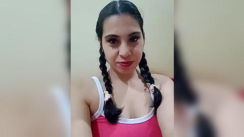 Media: Video of a young woman with light brown skin, long black hair in twin braids adorned with pink ribbons, wearing a red tank top with white trim. Background is a plain, light-colored wall.