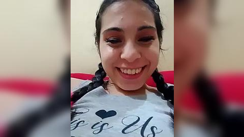 Media: Video of a smiling, light-skinned Latina woman with long dark hair in twin braids, wearing a gray t-shirt with black text. Background is a blurred, beige wall and red couch.