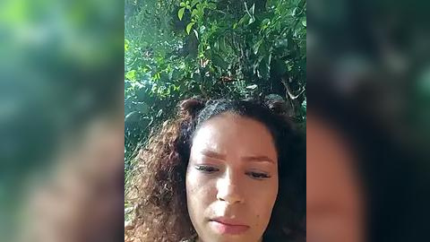 Media: Video of a young woman with curly brown hair, wearing a brown tank top, standing in a lush, green forest. Her expression is contemplative, surrounded by vibrant foliage.