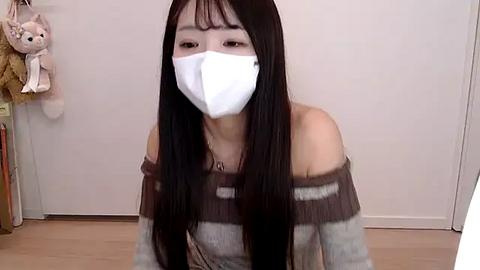 Media: A video of an Asian woman with long black hair, wearing a white face mask, off-shoulder gray sweater, and sitting on a wooden floor in a minimalist room with a beige wall and plush toys.