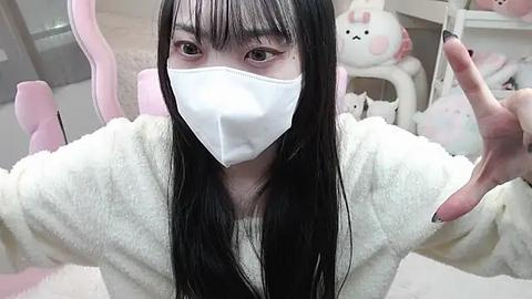 Media: Video of an Asian woman with long black hair, wearing a white mask, white fuzzy sweater, and pink nails. Background features pastel-colored furniture and plush toys.