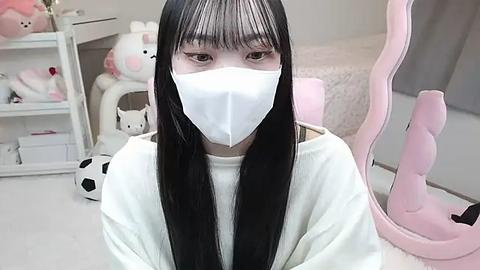 Media: Video of an Asian woman with long black hair and white mask, wearing a white sweater, seated in a pink-themed, plush-filled bedroom with white furniture and stuffed toys.