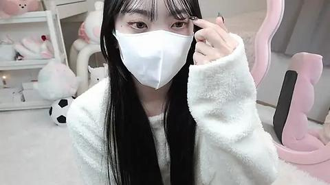 Media: Video of an Asian woman with long black hair, wearing a white mask, white fluffy robe, and pink hairbrush, in a plush, pastel-colored room with white shelves and stuffed animals.