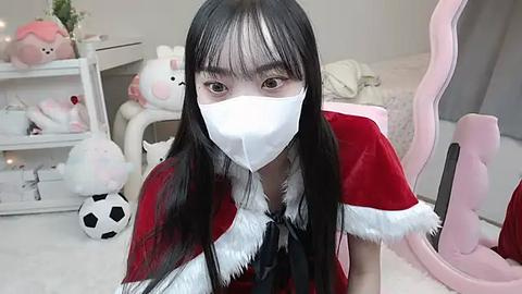 Media: A video of an Asian woman with long black hair, wearing a white mask and red Santa outfit, kneeling on a plush white carpet, surrounded by stuffed animals and a pink chair.