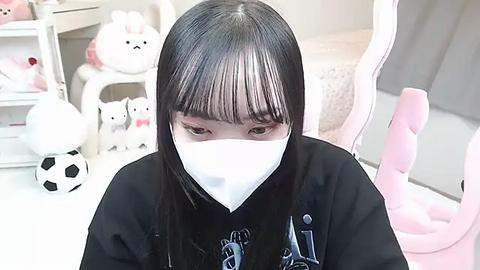 Media: Video of an Asian woman with long black hair and bangs, wearing a white mask, black hoodie, and seated in a pastel-colored gaming chair, surrounded by plush toys and a soccer ball.