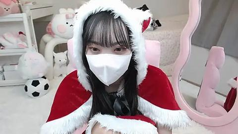 Media: Video of a young Asian woman with straight black hair, wearing a white face mask, red and white Santa hat, and a red velvet dress, seated in a pink gaming chair in a pastel-colored room filled with plush toys and a soccer ball.