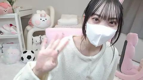 Media: Video of an Asian woman with long black hair and fair skin wearing a white face mask, seated in a pastel-colored gaming chair. Surroundings include plush toys and a white shelf.