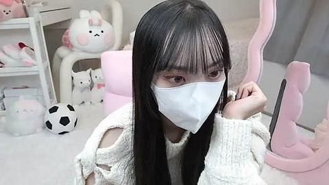 Media: Video of an East Asian woman with long black hair and fair skin, wearing a white face mask, a cream sweater, and a pink gaming headset, sitting on a pink chair in a cozy, pastel-themed room with plush toys.