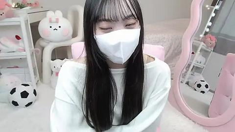 Media: Video of an East Asian woman with long black hair, wearing a white mask, white long-sleeve shirt, and a pink chair in a pastel-themed room with white walls, plush toys, and a pink mirror.