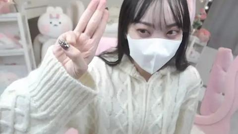 Media: Video of a young Asian woman with long black hair, wearing a white mask, white sweater, and pink chair, waving her left hand. Background includes white furniture and plush toys.