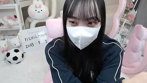 Media: A video of a young Asian woman with long black hair, wearing a white face mask and navy jacket, seated in a pink gaming chair, surrounded by plush toys and a soccer ball.