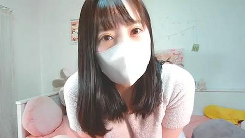 Media: Video of an Asian woman with long black hair, wearing a white mask, in a bedroom with white walls, a bed with pink and yellow pillows, and a framed photo.