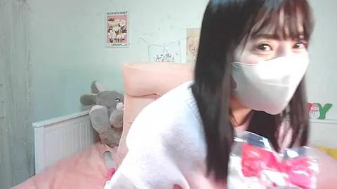 Media: Video of a young Asian woman with long black hair and fair skin, wearing a white mask, sitting on a pink bed with a plush gray bear, pastel wall with posters, and \"LOVE\" letters.