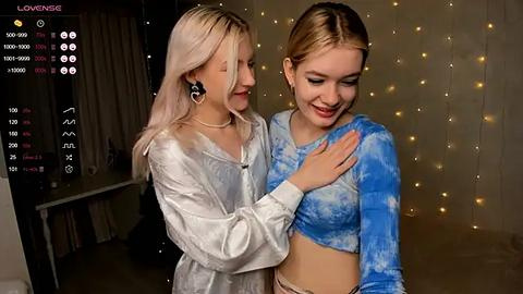 Media: Video of two young women, one in a white satin jacket and the other in a blue tie-dye crop top, smiling and hugging in a dimly-lit room with fairy lights.