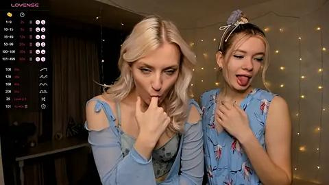 Media: Video of two young women with blonde hair, one licking fingers, wearing light blue floral dresses, standing against a backdrop of fairy lights in a dimly lit room.