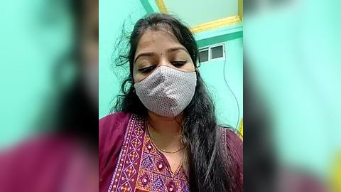Media: Video of a South Asian woman with long black hair, wearing a gray face mask and a maroon top with intricate embroidery, standing in a teal-painted room with a yellow ceiling.