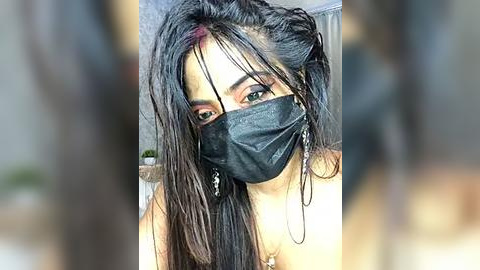 Media: Video of a young woman with long, dark, wet hair, wearing a black face mask, green eyes, and silver earrings, blurred background.