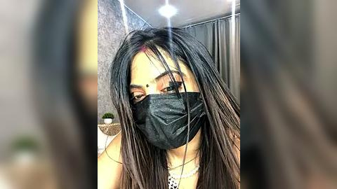 Media: Video of a young woman with long, straight black hair, wearing a black face mask and a silver necklace, in a dimly lit bathroom with a shower curtain.