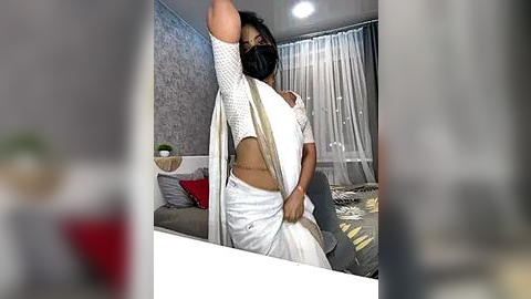 Media: Video of a South Asian woman with medium skin tone, wearing a white saree, black mask, and a white polka-dot blouse, stretching her arm above her head in a modern living room with gray wallpaper, white curtains, and a beige couch.