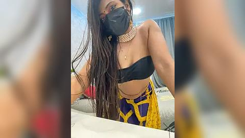 Media: Video of a woman with long, straight black hair, wearing a black face mask, black strapless top, and yellow and purple patterned pants, standing in a modern room with light walls and a bed.