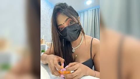 Media: Video of a South Asian woman with long, dark hair, wearing a black mask, and a black top, applying makeup in a modern, light-colored room with a blue wall and curtains.