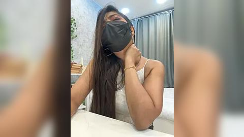 Media: Video of a woman with long brown hair, wearing a black face mask and a white tank top, leaning over a bathroom counter, looking distressed.