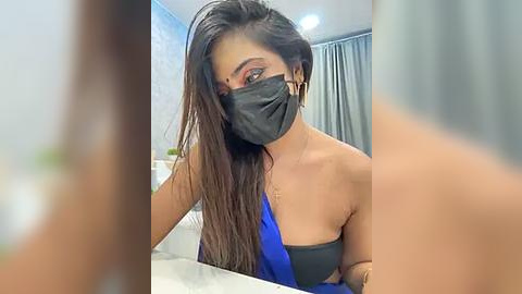 Media: Video of a young woman with long dark hair, wearing a black face mask and a blue strapless top, sitting in a modern room with light walls and a grey curtain.