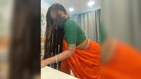 Media: Video of a South Asian woman in a green blouse and orange saree, bending over a table, partially obscured by another person.