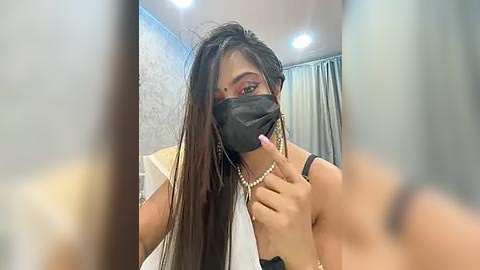 Media: A video shows a woman with long, dark hair, wearing a black mask and a white top, touching her mouth with a finger. She's in a room with a grey wall and a white curtain.