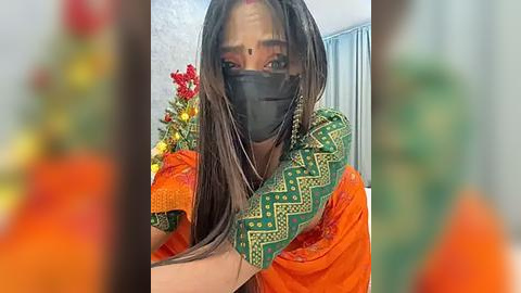 Media: A video of a South Asian woman with medium skin tone, wearing a green sari with orange border, black mask, and long dark hair. Background includes a blurred floral arrangement and light blue curtains.