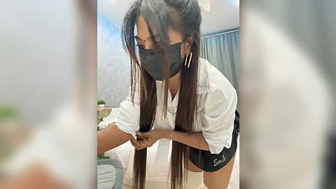 Media: Video of an Asian woman with long hair, wearing a black mask, white shirt, and black shorts, adjusting her hair in a bedroom with light gray walls.