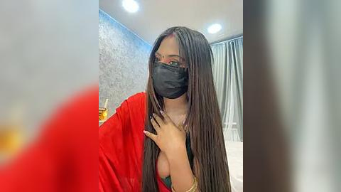 Media: Video of a woman with long black braids, wearing a red saree, black face mask, and black top, indoors with grey textured wall and white curtains.