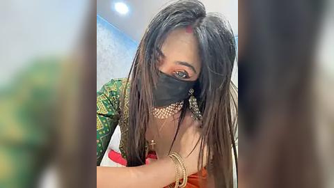 Media: Video of a South Asian woman with long, straight black hair and a black mask covering her nose and mouth, wearing a green saree with gold embroidery and multiple necklaces.