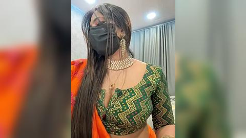 Media: Video of a South Asian woman in a green and gold traditional blouse with a black mask, black face veil, and large gold earrings, standing indoors with soft lighting.