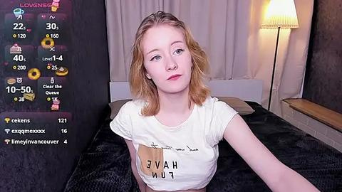 Media: Video of a young woman with fair skin and blonde hair, wearing a white T-shirt with a \"JAV\" logo, sitting on a bed with a dark bedspread, in a dimly lit room.