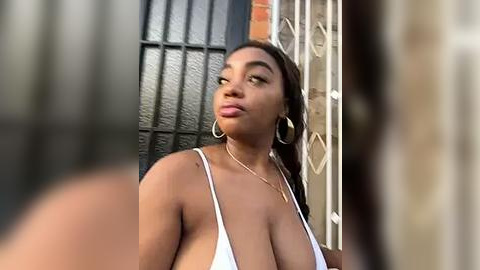 Media: Video of a curvy, light-skinned Black woman with long dark hair, wearing a white bikini, looking confident outdoors near a metal gate and brick wall.