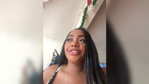 Media: A video of a smiling Black woman with long, straightened hair, wearing a patterned top, standing indoors with festive green garland and red ornaments hanging above her.