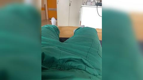 Media: Video of a person in green scrubs lying on a bed with white sheets, blurred background showing a wooden desk and wall.