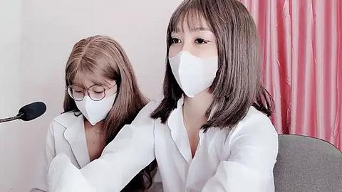 Media: Video of two young women with straight, shoulder-length brown hair, wearing white lab coats and face masks, seated in a lab setting with pink curtains and a microphone.