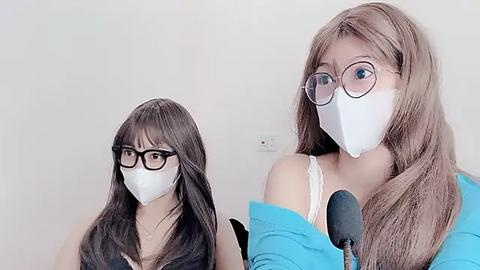 Media: Video of two East Asian women with long, straight brown hair, wearing white face masks and black-rimmed glasses, sitting close together against a plain white wall.