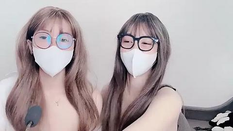 Media: Video of two Asian women with long, straight brown hair, wearing white face masks and glasses, smiling, seated indoors, against a plain white background.