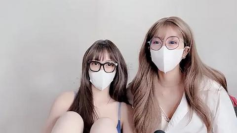 Media: Video of two young women with long hair, wearing white face masks and oversized glasses, sitting against a plain white background, in casual attire.