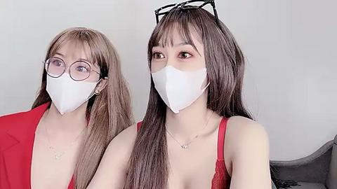 Media: Video of two East Asian women wearing white masks, glasses, and red tops. One has long, straight hair, while the other has short, wavy hair. Background is a plain, light-colored wall.