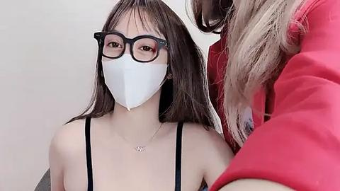 Media: Video of a young Asian woman with straight black hair, wearing black-framed glasses, a white surgical mask, and a black spaghetti strap top, with another person's blonde hair visible in the background.