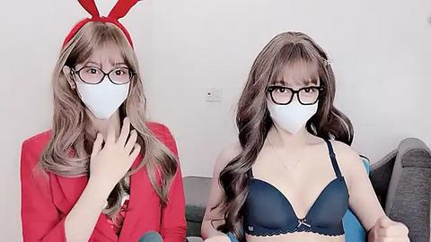 Media: Video of two women in matching white masks, one in a red dress, the other in a blue bra, both with long, wavy hair and glasses, sitting side-by-side on a couch.